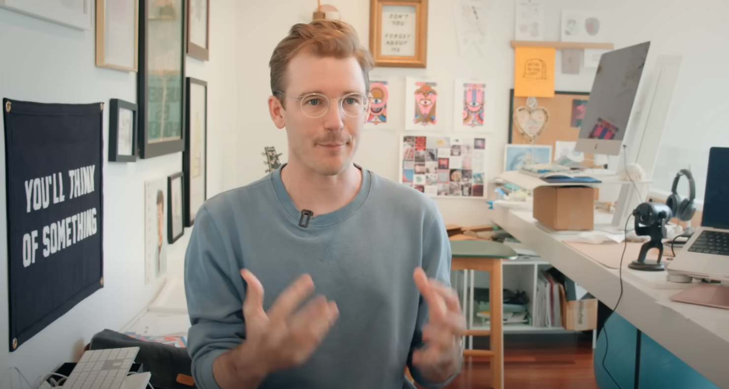 a still from the React documentary showing Lee sitting at a desk in front of a large black cloth banner embroidered with the words "You'll Think of Something" in white block letters